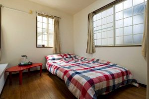 Habitación con suelo de madera, mesita roja con kit de té, cama en la esquina con colcha cuadros de rayas azules y rojas, dos ventanas grandes, pared blanca.