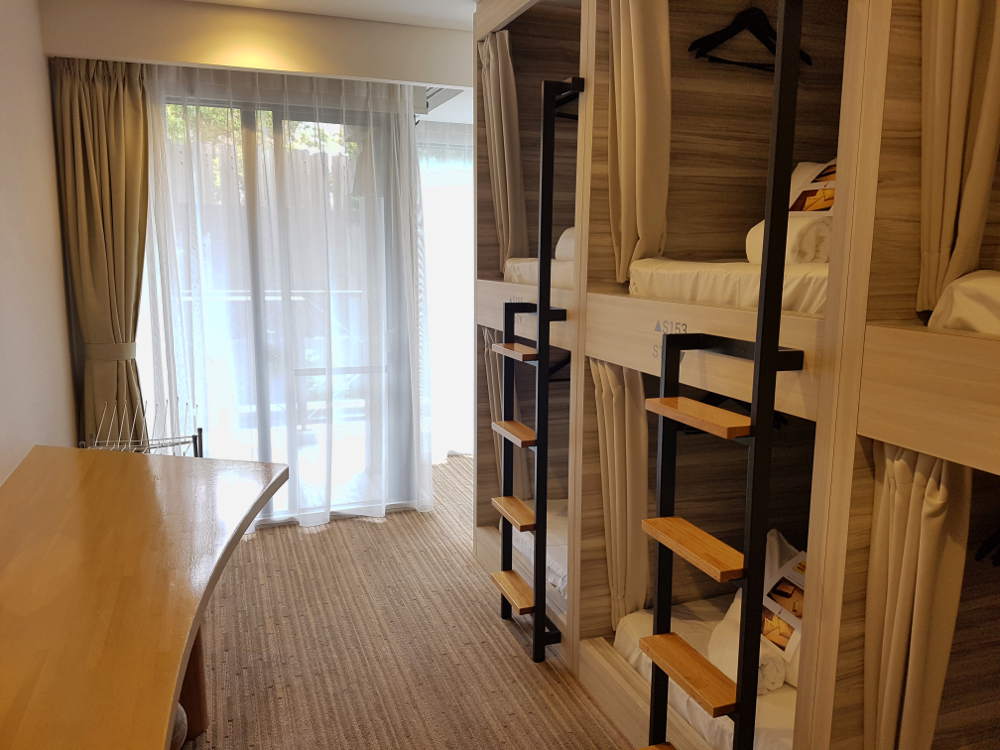 buscar alojamiento en Japón: Habitación con moqueta a rayas marrones y ventanal al fondo con cortina blanca. A la derecha literas dobles de cerradas de madera.