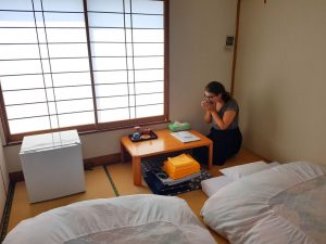 Habitación tradicional japonesa, suelo de bambú, ventanas de madera y papel blanco traslúcido, fotones blancos, nevera pequeña y mesita con bandeja de té. Carla de rodillas con ambas manos coge un vaso de té y se lo lleva a la boca.