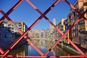 En primer plano, reja de acero en forma de cruz en color rojo y, de fondo, la ciudad de Girona con el río en medio.