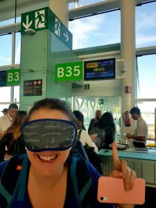 primer plano de carla sonriente con antifaz cubriendo sus ojos, de fondo la puerta de embarque del aeropuerto con panel que pone destino ibiza, ventanales con aeropuerto de fondo y cielo despejado
